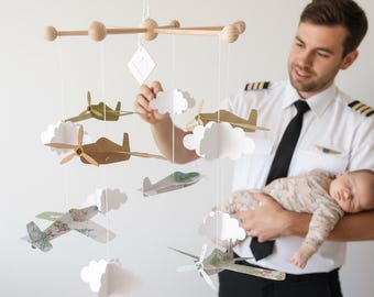 May include: A wooden baby mobile featuring paper airplanes and white paper clouds suspended from a wooden frame. The airplanes are in various colors and patterns. The mobile is designed for a nursery.