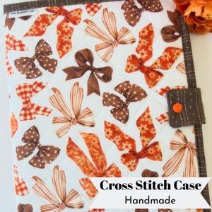 May include: A quilted fabric case featuring a white background with a repeating pattern of brown and orange bows in various styles, including polka dot, striped, and floral prints. It has a dark brown border and a snap closure with an orange button.