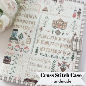 May include: A quilted fabric case featuring an off-white background with intricate cross-stitch patterns including alphabets, numbers, houses, trees, and figures. The case has a brown and white striped border and a snap button closure.