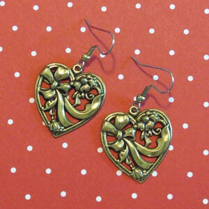 May include: A pair of gold heart-shaped earrings with a delicate floral design. The earrings feature a bow-like detail and hang from silver hooks.