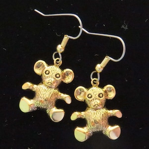 May include: A pair of gold-tone earrings featuring a teddy bear charm. The teddy bears have a textured surface and are wearing a simple, gold-tone earring hook.