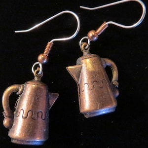 Peut inclure: Une paire de boucles d'oreilles en cuivre en forme de cafetières avec un crochet argenté. Les cafetières ont un motif ondulé autour du bas.