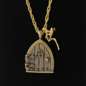 Könnte beinhalten: Eine goldene Kettenkette mit einem bronzenen Anhänger in Form einer kleinen Tür mit einem Fenster und einer kleinen Feenfigur, die oben befestigt ist.