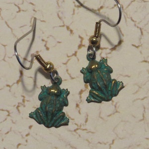 Puede incluir: Un par de pendientes de rana verde con detalles dorados y ganchos plateados.
