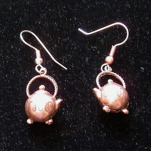 May include: A pair of silver dangle earrings with a copper teapot charm. The teapot charm has a small spout and handle and is engraved with the word "CUP".