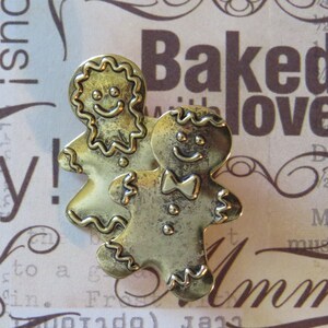May include: Two gold-coloured gingerbread man charms with smiling faces. The charms are detailed with raised lines and have a slightly worn look.