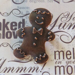 May include: A copper-coloured gingerbread man charm with a wavy outline and a simple bow tie. The charm has a smiling face and two small dots for eyes.