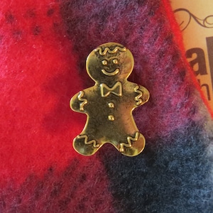 May include: A gold-coloured gingerbread man ornament with a bow tie and a smiling face. The ornament is on a red and black fuzzy fabric.
