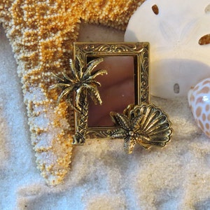 Puede incluir: Un marco de fotos dorado con un diseño de palmera y concha. El marco está sobre una superficie de arena blanca con una estrella de mar y conchas.