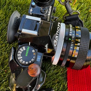 May include: Black Canon camera with a lens, strap, and a wooden dial. The lens has distance markings in feet and meters. The camera is on a bed of green grass with a red strap.