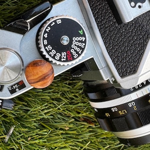 May include: A silver and black camera with a wooden knob and a black lens. The camera has a dial with numbers and letters, including "B", "1", "2.5", "4", "8", "15", "30", "60", "125", "250", "500", "1000", "2000", "4000". The lens has a depth of field scale with numbers from 0.5 to 15.