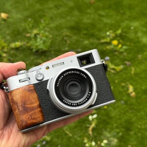 May include: A silver and black digital camera with a wooden grip is held in a hand. The lens is black with white text that reads "FUJINON ASPHERICAL LENS SUPER EBC f=23mm 1:2". The camera is on a green grassy background.