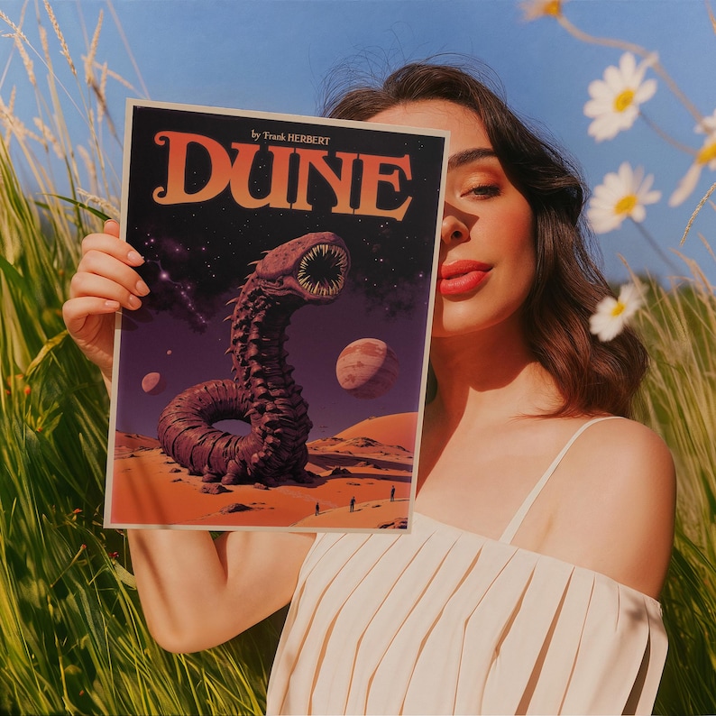 Może przedstawiać: Kobieta trzyma okładkę książki przedstawiającą piaskowego robaka z powieści science fiction "Diuna" Franka Herberta. Okładka jest ilustrowana ciemnofioletowym, czerwonym i pomarańczowym piaskowym robakiem na tle gwieździstego nocnego nieba.