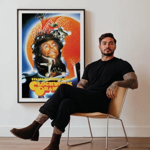 May include: A framed poster for Stanley Kubrick's "A Clockwork Orange" with a surreal image of a man's face inside an orange. The poster is in a black frame. A person is sitting in a chair in front of the poster.