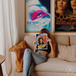 May include: A person seated on a beige sofa holds a framed movie poster for "A Clockwork Orange", with a vibrant orange and yellow background. Two other framed movie posters are visible: "Blue Velvet" and "Mulholland Drive". The room has a neutral colour palette.