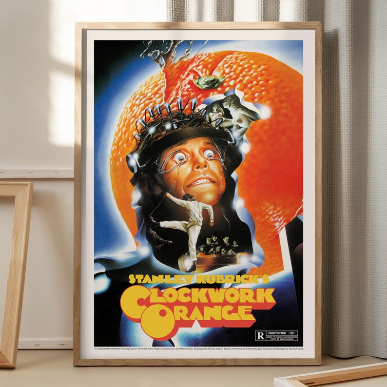 May include: Framed poster for Stanley Kubrick's "A Clockwork Orange." The artwork features a man with a headpiece, set against an orange and blue backdrop. The film's title is in orange and yellow. The poster is in a wooden frame.