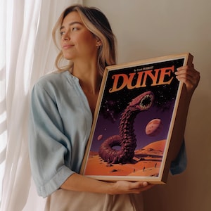 Może przedstawiać: Ramka z plakatem w stylu retro z tytułem "Diuna" autorstwa Franka Herberta. Plakat przedstawia duże stworzenie przypominające piaskowego robaka z otwartą paszczą, na tle gwieździstego nieba i pustynnego krajobrazu.