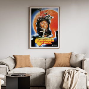 May include: A framed poster for "A Clockwork Orange" is displayed above a light-coloured sofa with two brown cushions. The poster features a portrait of a man with orange and blue accents. The room has wooden shelves and a black side table.