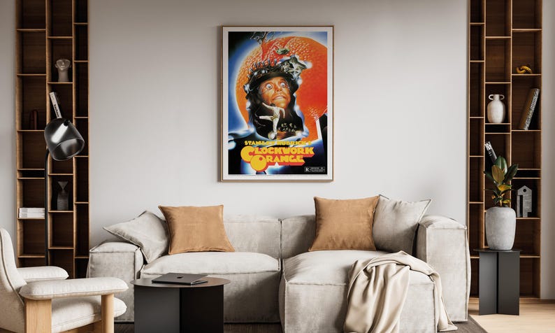 May include: A framed poster for "A Clockwork Orange" is displayed above a light-coloured sofa with two brown cushions. The poster features a portrait of a man with orange and blue accents. The room has wooden shelves and a black side table.