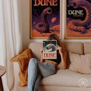 Może przedstawiać: Kobieta siedzi na kanapie trzymając okładkę książki z piaskowym robakiem i tekstem "Dune" w czerwonych literach. Okładka książki jest przed jej twarzą. Dwa plakaty z okładką książki znajdują się na ścianie za nią.