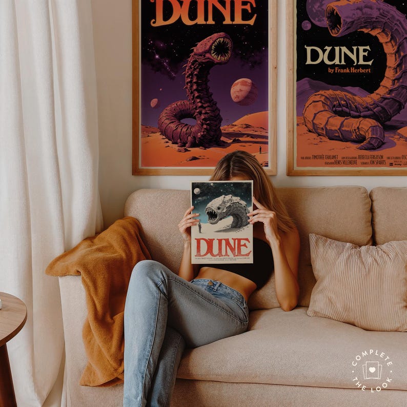 Może przedstawiać: Kobieta siedzi na kanapie trzymając okładkę książki z piaskowym robakiem i tekstem "Dune" w czerwonych literach. Okładka książki jest przed jej twarzą. Dwa plakaty z okładką książki znajdują się na ścianie za nią.