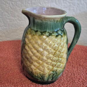 Peut inclure: Pichet en céramique avec un motif d'ananas. Le pichet a une anse et un bord verts, avec un corps texturé jaune et crème. Il est probablement utilisé pour servir des boissons.