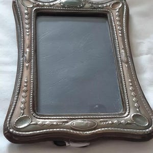 May include: A silver-toned picture frame with a decorative border and a dark grey mat. The frame has a wavy, ornate design with oval-shaped accents.