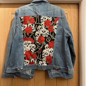 May include: Pale blue denim jacket with a black panel on the back. The panel displays a pattern of white skulls and red roses. The jacket has a classic design with button closures and pockets.