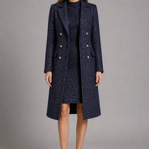 Op de afbeelding: Een marineblauwe tweed jas met gouden knopen, gedragen over een bijpassende jurk. De jas is dubbelknoops en knielang. De jurk heeft een hoge halslijn. De outfit wordt gecompleteerd met zwarte hakken.