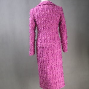 May include: A pink skirt suit with a textured, fringed fabric. The jacket has a collar and long sleeves. The skirt is knee-length.