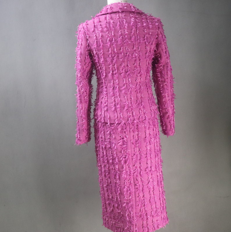 May include: A pink skirt suit with a textured, fringed fabric. The jacket has a collar and long sleeves. The skirt is knee-length.