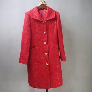 May include: A red tweed coat with a classic collar and three gold buttons. The coat has two front pockets and is embellished with small, sparkling sequins. The coat is hanging on a wooden hanger.