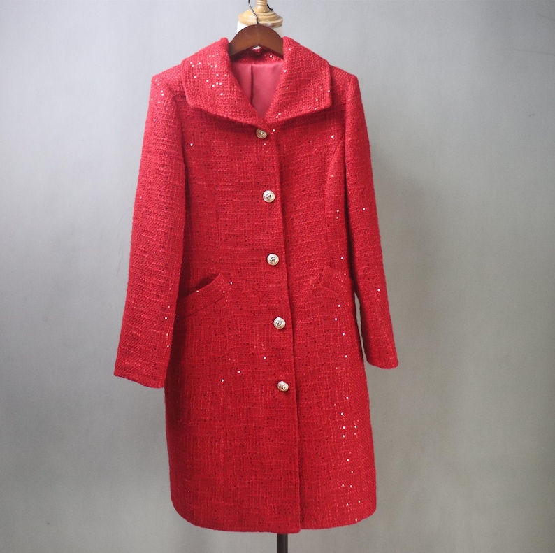May include: A red tweed coat with a classic collar and three gold buttons. The coat has two front pockets and is embellished with small, sparkling sequins. The coat is hanging on a wooden hanger.
