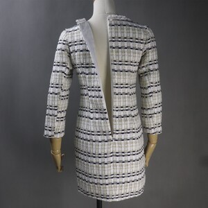 May include: A tweed dress with a cream, black, and tan checkered pattern. The dress has long sleeves and a zipper down the back. The dress is displayed on a mannequin.