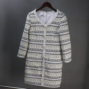May include: A long-sleeved, tweed dress in a cream, black, and gold plaid pattern. The dress features a V-neckline, decorative buttons, and fringe trim along the collar and pockets. The dress is hanging on a wooden hanger.