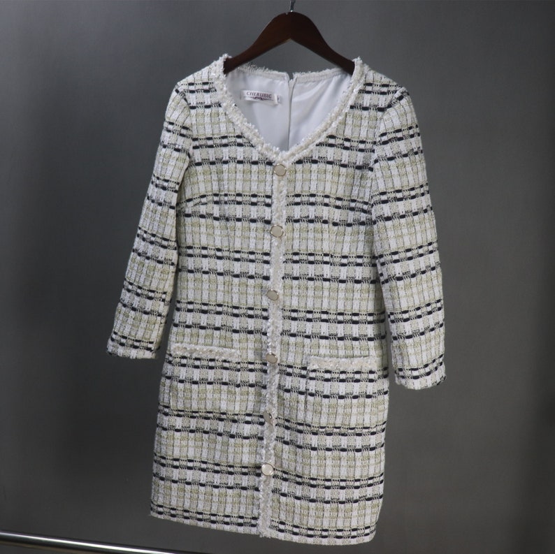 May include: A long-sleeved, tweed dress in a cream, black, and gold plaid pattern. The dress features a V-neckline, decorative buttons, and fringe trim along the collar and pockets. The dress is hanging on a wooden hanger.
