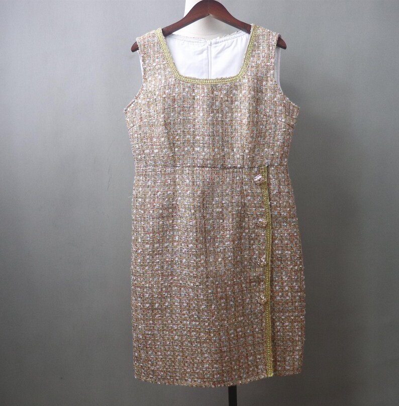 May include: Sleeveless tweed dress in a neutral color palette of beige, white, and gold. The dress features a square neckline, a fitted waist, and a straight skirt. Gold trim accents the neckline and the front closure. The dress is hanging on a wooden hanger.