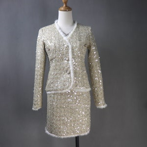 Può includere: Un completo giacca e gonna in paillettes beige e bianco. La giacca ha un collo a V e un bordo in pelliccia bianca. La gonna è lunga fino al ginocchio e ha anche un bordo in pelliccia bianca.