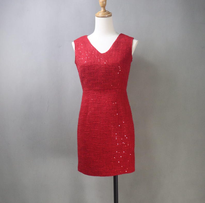 May include: A red, sleeveless dress with a V-neckline. The dress is made of a textured fabric and is adorned with scattered sequins. The dress is displayed on a mannequin.