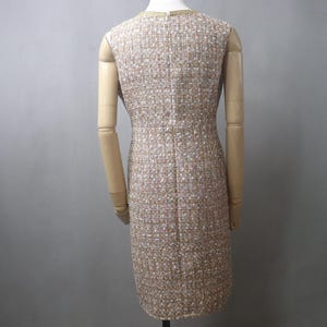May include: A sleeveless tweed dress in a neutral color palette of beige, white, and gold. The dress has a sheath silhouette and a subtle textured pattern. The neckline is trimmed with gold detailing. The dress is displayed on a mannequin.