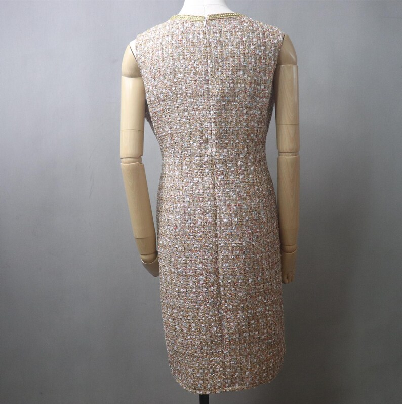 May include: A sleeveless tweed dress in a neutral color palette of beige, white, and gold. The dress has a sheath silhouette and a subtle textured pattern. The neckline is trimmed with gold detailing. The dress is displayed on a mannequin.