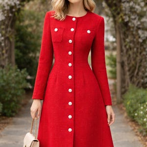 May include: A red, long-sleeved dress with a button-down front and a-line skirt. The dress features white buttons and small pockets. A small, beige purse with a gold chain is held in the hand.