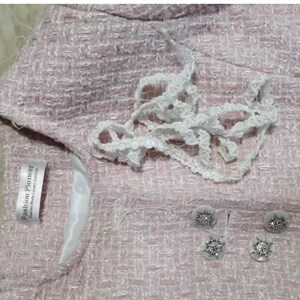 May include: Close-up of a light pink tweed jacket with white lace trim and decorative buttons. The jacket has a label that reads "Fashion Pioneer". The buttons are silver with a floral design.