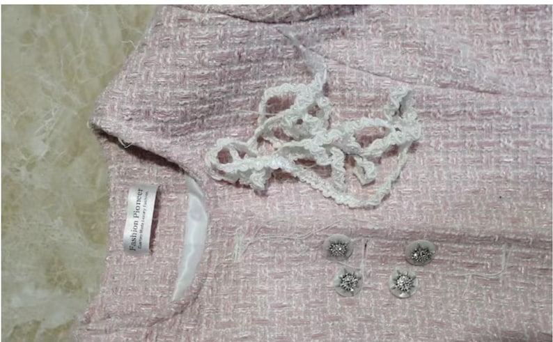 May include: Close-up of a light pink tweed jacket with white lace trim and decorative buttons. The jacket has a label that reads "Fashion Pioneer". The buttons are silver with a floral design.
