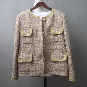 May include: A tan tweed jacket with gold trim around the collar, pockets, and cuffs. The jacket has a round neckline, four front pockets, and a button-up closure. The fabric has a woven texture with flecks of white and brown.