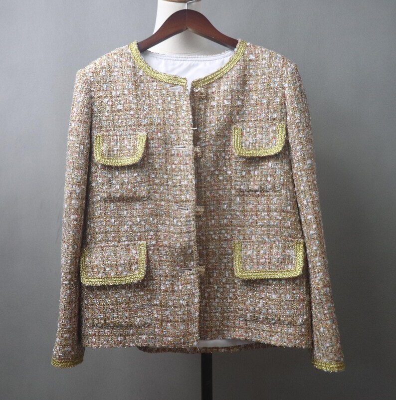 May include: A tan tweed jacket with gold trim around the collar, pockets, and cuffs. The jacket has a round neckline, four front pockets, and a button-up closure. The fabric has a woven texture with flecks of white and brown.