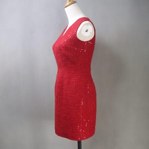 May include: A red, textured, V-neck dress with subtle sequin accents. The dress is displayed on a mannequin, showcasing its fitted silhouette and knee-length hemline. The fabric appears to have a woven texture, adding depth to the design.