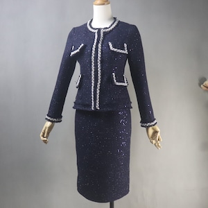 May include: A navy blue tweed skirt suit. The jacket has a round neckline, long sleeves, and front pockets. The edges of the jacket, pockets, and sleeves are trimmed with white beading. The skirt is knee-length. The suit is displayed on a mannequin.