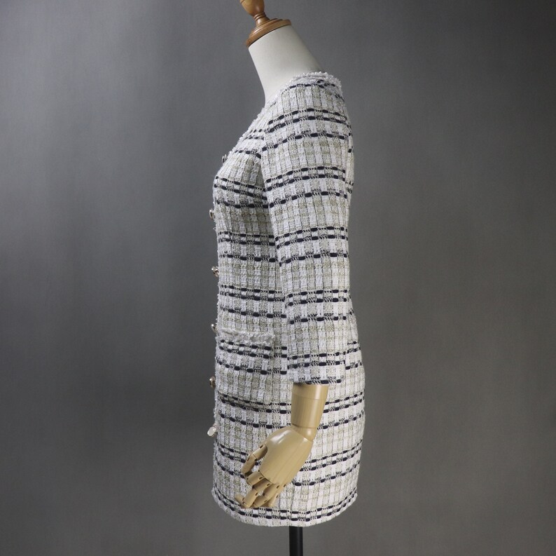 May include: A cream-colored tweed dress with a checkered pattern in black and beige. The dress features long sleeves, gold buttons, and two front pockets. The dress is displayed on a mannequin.