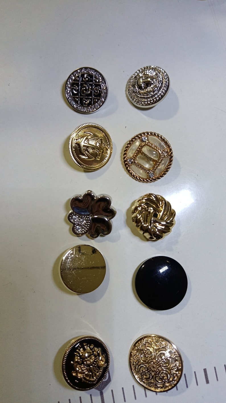 May include: A collection of decorative buttons in various designs and colors. Includes gold, silver, and black buttons with intricate patterns, including floral, geometric, and nautical motifs. Some buttons have rhinestones.